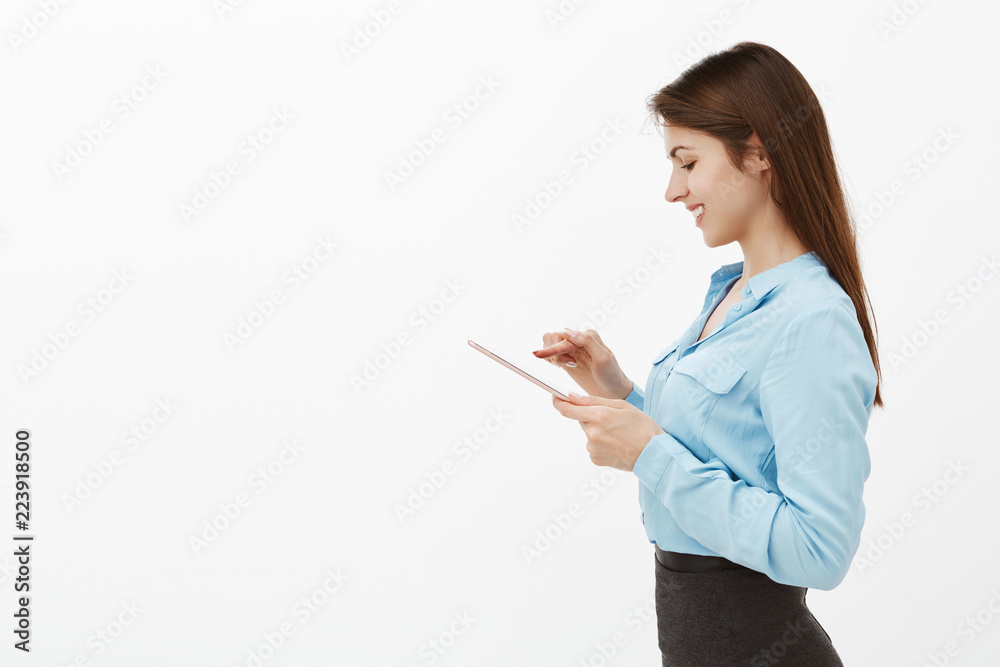Positive good-looking entrepreneur in blue blouse, typing message in digital tablet, smiling broadly at screen, talking with friend or coworker from another department, standing over grey wall Positive good-looking entrepreneur in blue blouse, typing message in digital tablet, smiling broadly at screen, talking with friend or coworker from another department, standing over grey wall