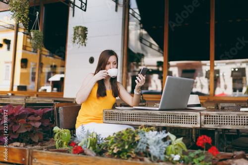 Happy girl in outdoors street coffee shop cafe sitting at table with laptop pc computer, texting message on mobile phone, drink cup tea in restaurant during free time. Mobile office freelance concept.