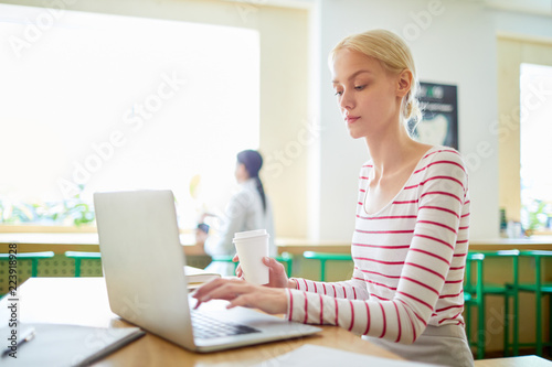 Young woman with drink sitting by table in front of laptop and communicating with someone