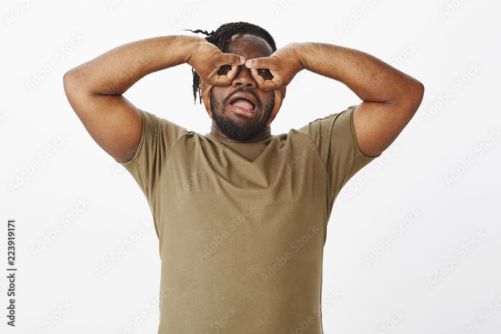Waist-up shot of funny carefree african-american guy in olive t-shirt, holding hands over eyes and making faces, mimicking goggles or eyewear while fooling around over grey background Waist-up shot of funny carefree african-american guy in olive t-shirt, holding hands over eyes and making faces, mimicking goggles or eyewear while fooling around over grey background
