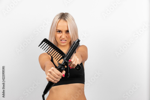 A girl with blond hair holds a black large plastic comb and a black hair dryer and is angry against a white background