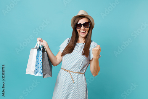 Portrait fashionable attractive happy woman in summer dress, straw hat, sunglasses holding packages bags with purchases after shopping isolated on blue pastel background. Copy space for advertisement.