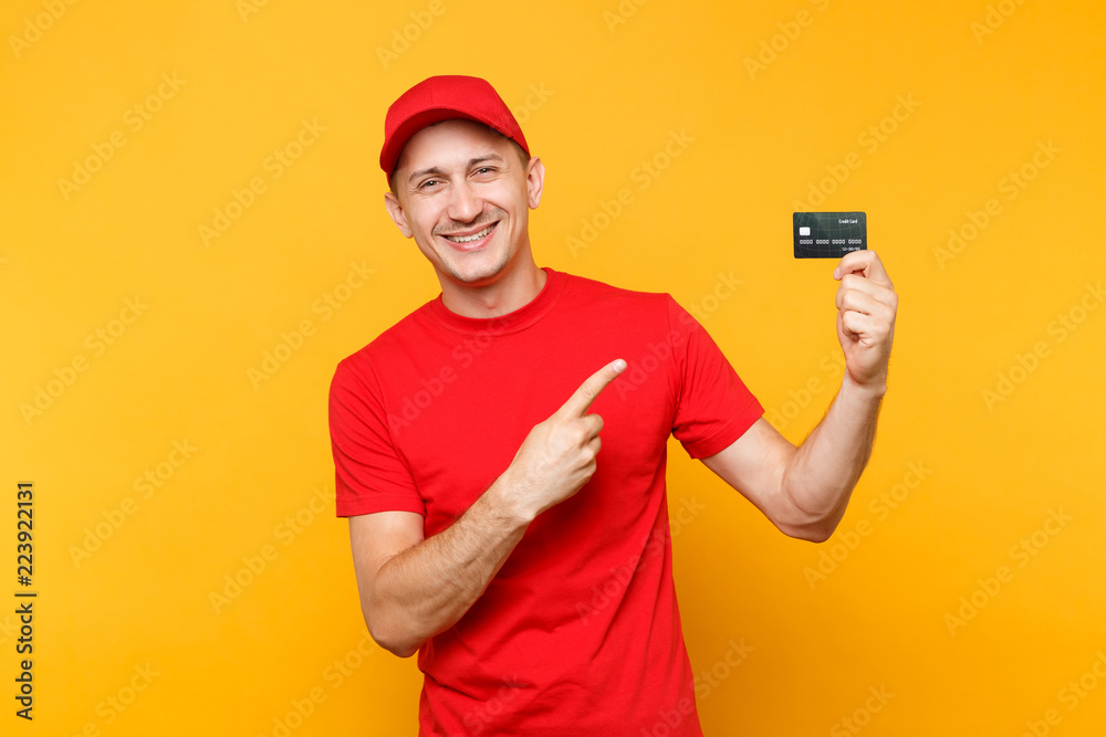 Delivery man in red uniform isolated on yellow orange background. Professional smiling male employee in cap, empty t-shirt courier or dealer hold in hand credit bank card. Service concept. Copy space. Delivery man in red uniform isolated on yellow orange background. Professional smiling male employee in cap, empty t-shirt courier or dealer hold in hand credit bank card. Service concept. Copy space.