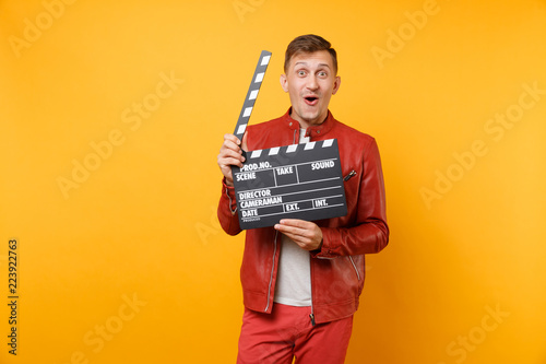 Portrait vogue smiling young man in red leather jacket, t-shirt holding classic black film making clapperboard isolated on bright trending yellow background. People lifestyle concept. Advertising area
