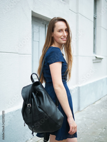 Hipster young redhead girl with freckles wearing blue dress and black backpack posing against city street building wall
