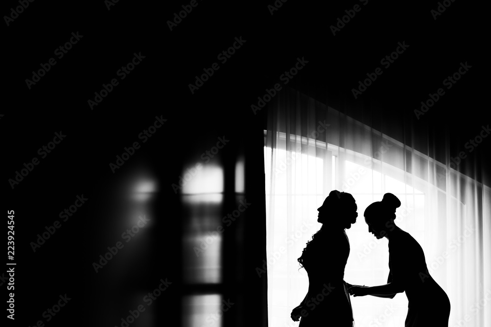 Profile of the bride and her friend who lacing corset in a wedding dress near the window on a black and white photo