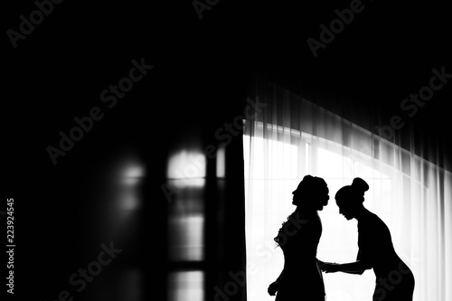 Profile of the bride and her friend who lacing corset in a wedding dress near the window on a black and white photo