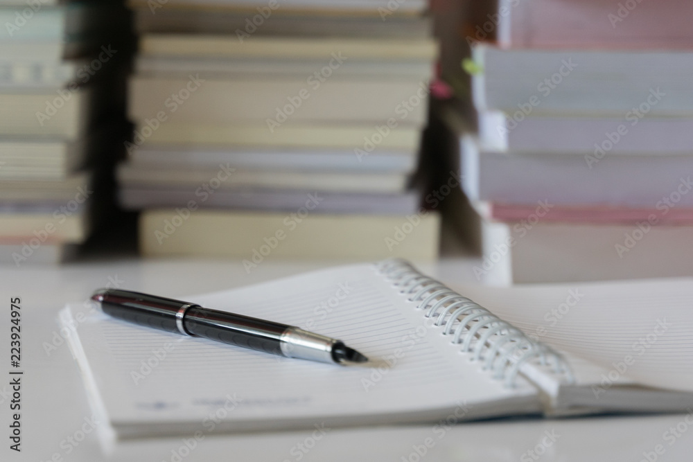 Close up of notebook with ink-pen and eyeglasses put on white table with blur focus of books as a background. Educational concept. Close up of notebook with ink-pen and eyeglasses put on white table with blur focus of books as a background. Educational concept.