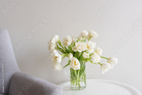 Close up of white double tulips in glass jar on small white table next to retro grey chair (selective focus)
