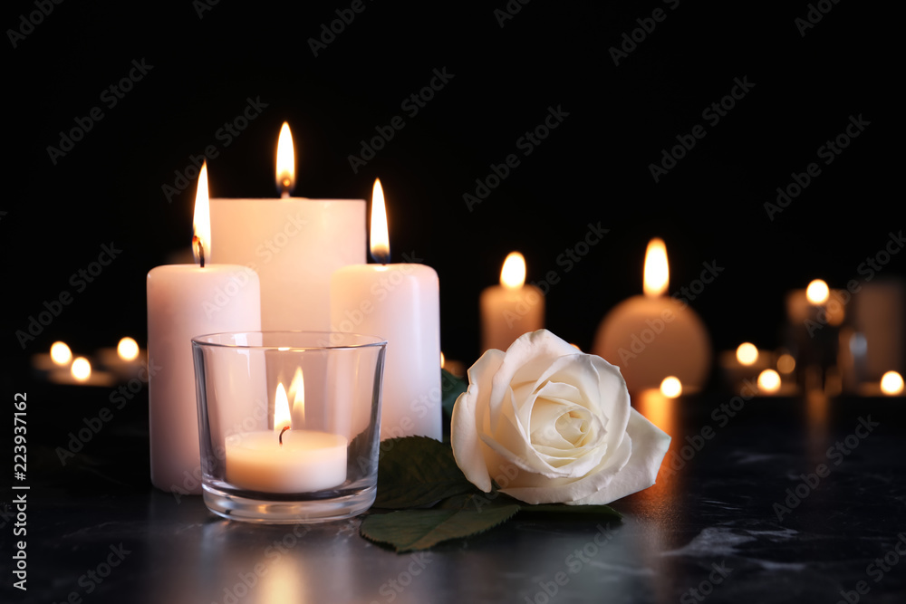 Fototapeta premium Biała róża i płonące świeczki na stole w ciemności. Symbol pogrzebu