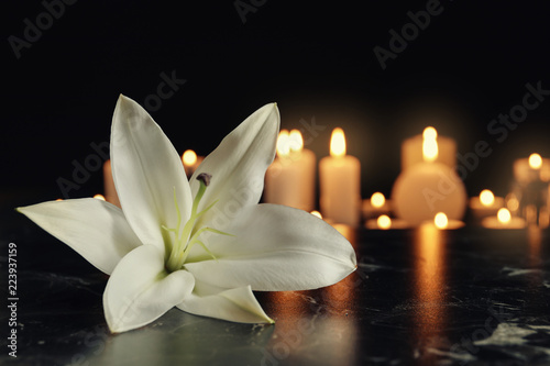 Fototapeta Naklejka Na Ścianę i Meble -  White lily and blurred burning candles on table in darkness, space for text. Funeral symbol