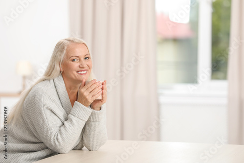 Portrait of beautiful older woman with cup of tea sitting at table indoors. Space for text