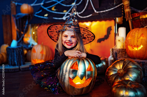 Happy Halloween. A little beautiful girl in a witch costume celebrates at home in an interior with pumpkins and cardboard magic house on the background