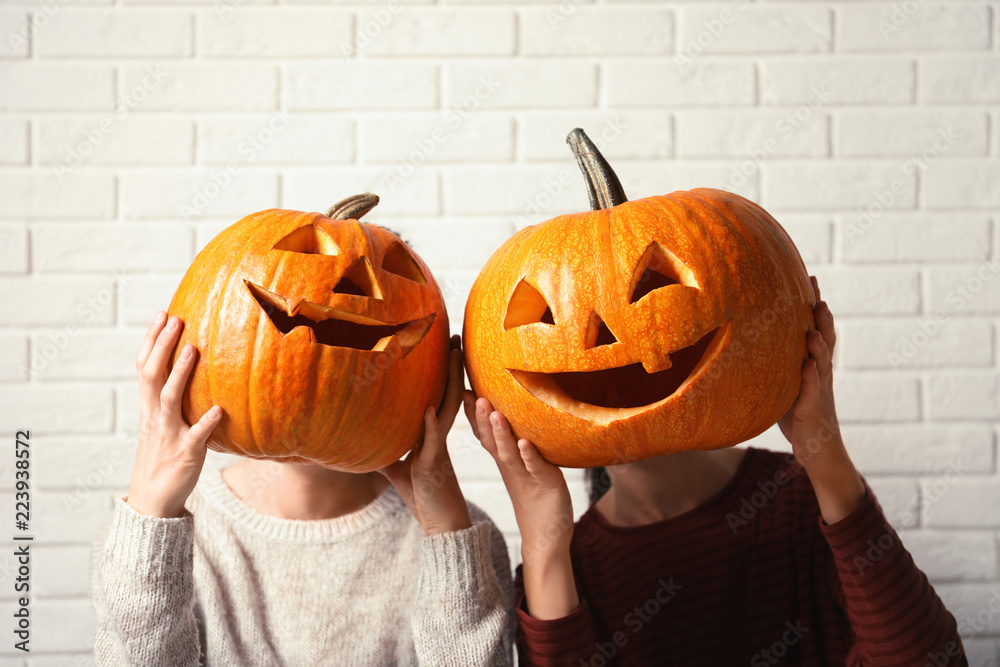 Fototapeta premium Women holding Halloween pumpkin head jack lanterns against brick wall
