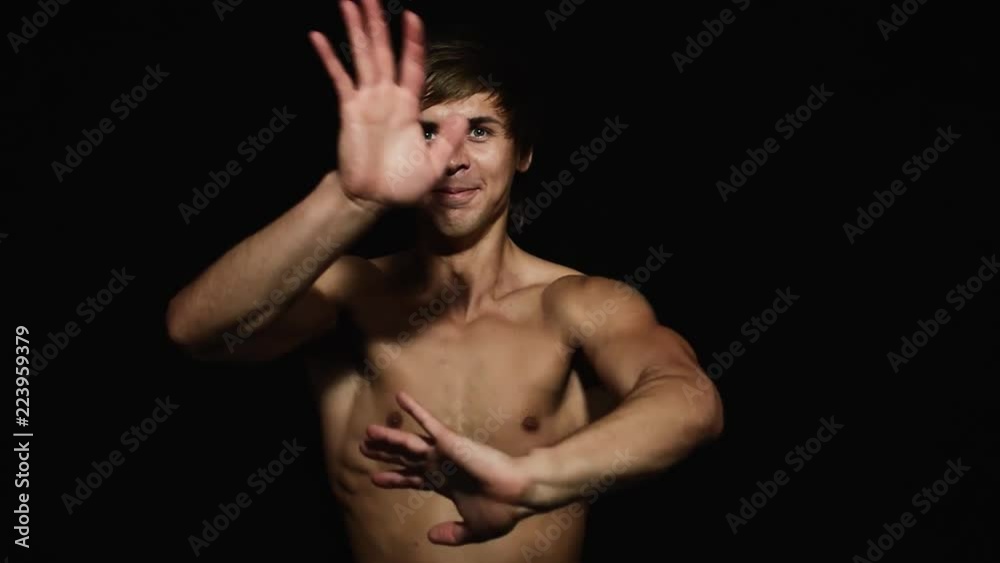 Young athletic attractive blue eyed male with naked torso and having modern haircut is smiling and dynamically waving his hands in front of camera on black matte background. Young athletic attractive blue eyed male with naked torso and having modern haircut is smiling and dynamically waving his hands in front of camera on black matte background.