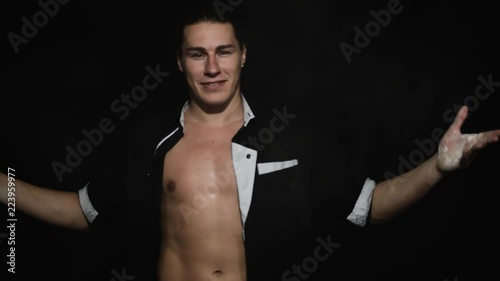 Handsome athletic young brown eyed guy with unbuttoned shirt having hair combed at nape is smiling and dynamically clapping with hands covered by white powder in front of camera on black background. photo