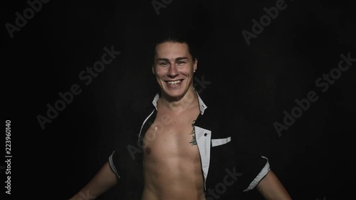 Handsome athletic young brown eyed male in unbuttoned shirt with tattoo on chest is smiling and throwing white powder on himself in front of camera on black matte background. photo