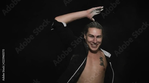 Handsome athletic young brown eyed male in unbuttoned shirt with tattoo on chest is smiling and playfully throwing white powder around himself in front of camera on black matte background. photo