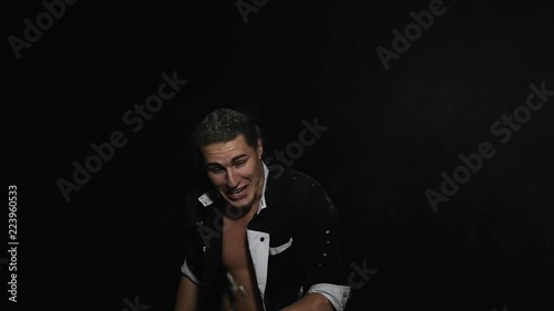White powder is being thrown on handsome athletic young brown eyed man with hair combed into bundle and dressed in unbuttoned shirt with tattoo on chest in front of camera on black matte background. photo