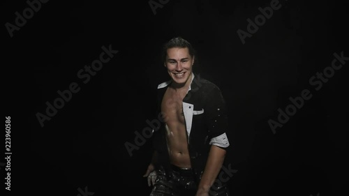 White powder is being thrown on handsome, sporty young brown eyed man with dark hair combed into bundle and wearing unbuttoned shirt with tattoo on chest in front of camera on black matte background photo