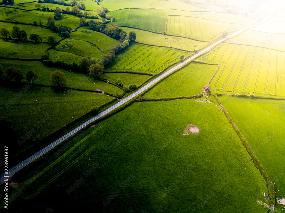 Aerial view looking down on a rural road in the UK countryside. On a bright sunny day, farmland and crops can be seen either side of the road