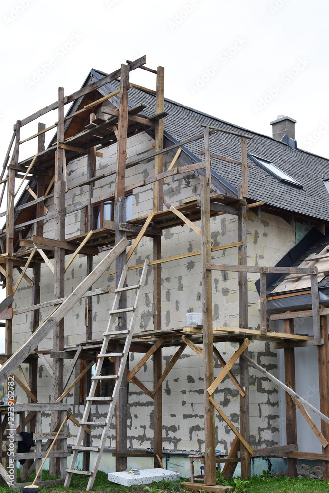 Wooden scaffolding next to white residental house built of aerated concrete blocks  with grey asphalt shingles roof on sky background with copy space. House improvement process nd DIY concept.
