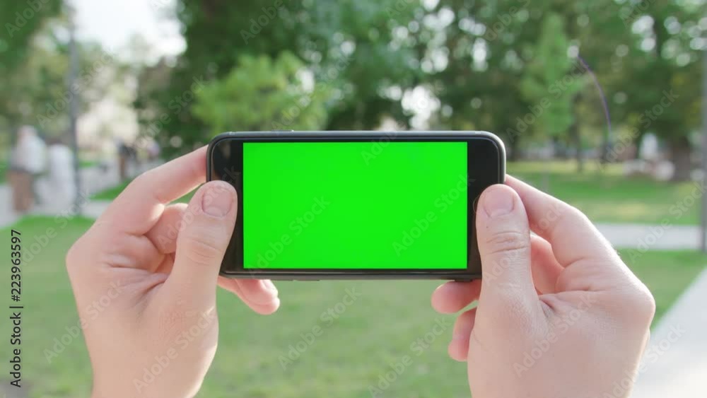 Two hands holding a phone with a green screen in a horizontal position in a public park. Close-up shot. Soft focus Two hands holding a phone with a green screen in a horizontal position in a public park. Close-up shot. Soft focus