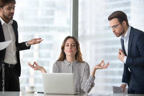 Calm female worker meditate at workplace managing stress not paying attention to angry colleagues, businesswoman sitting in lotus pose practicing yoga staying calm not involved in conflicts or dispute photo