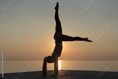 Side view of silhouette of a woman  standing on her hands. Healthy flexible girl doing yoga on sunrise. Girl practicing the handstand pose on tranquil beach at sunset during summer vacation in Spain.