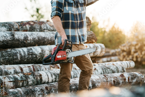 Professional bearded lumberman wearing plaid shirt hold chainsaw in hand on background of sawmill and warehouse of trees