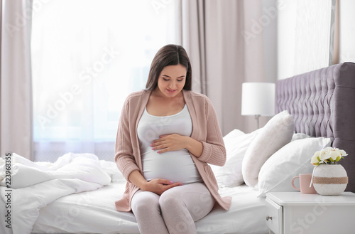 Young beautiful pregnant woman sitting on bed and touching her belly at home