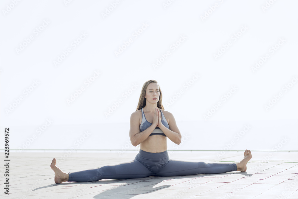 Fototapeta premium Beautiful girl with long hair dressed in sportswear doing the splits in urban exterior. Flexible woman doing yoga meditation with the white sky on the background. Calmness, relax, healthy lifestyle