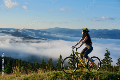Young female rider riding on bicycle in the mountains, wearing helmet, on summer morning. Foggy mountains, forests on the blurred background. Outdoor sport activity, lifestyle concept. Copy space