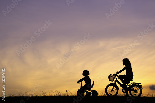 The silhouette photo of mother and her daughter riding bicycle in the park with sweet sky at sunset.