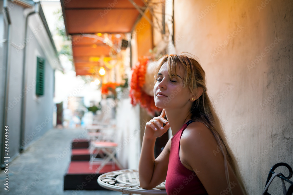 Beautiful tourist woman in local restaurant waiting for food sitting outdoor on mediterranean narrow street, travel lifestyle