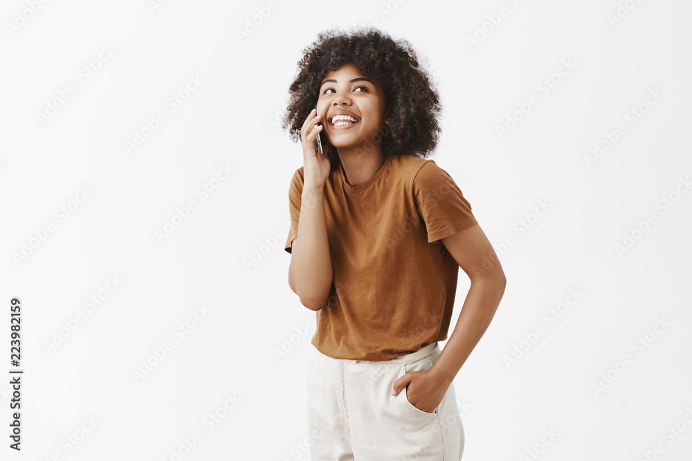 Studio shot of relaxed African American girl having fun talking casually with friend via smarpthone gazing up with carefre joyful smile holding hand in pocket standing over gray background Studio shot of relaxed African American girl having fun talking casually with friend via smarpthone gazing up with carefre joyful smile holding hand in pocket standing over gray background
