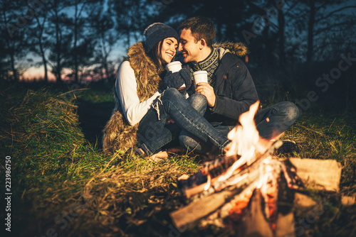 Pretty young couple drinking hot beverage in the forest near bonfire.