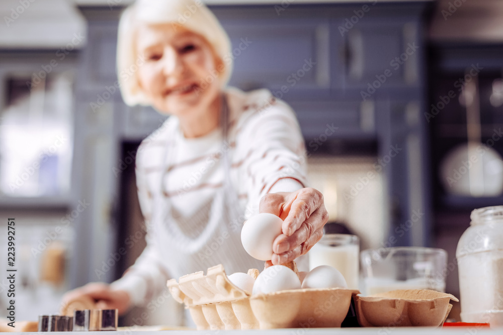 Egg for meal. Peaceful senior woman standing in the modern kitchen and carefully taking one egg for her meal