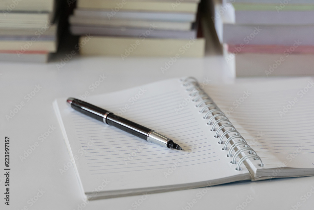Close up of notebook with ink-pen and eyeglasses put on white table with blur focus of books as a background. Educational concept. Close up of notebook with ink-pen and eyeglasses put on white table with blur focus of books as a background. Educational concept.
