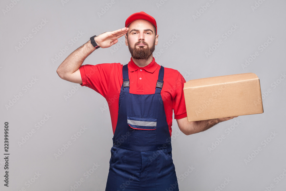 Give salute or yes sir! Young confident logistic delivery man with beard in blue uniform and red t-shirt standing, holding cardboard box on grey background. Indoor, studio shot, isolated, copy space Give salute or yes sir! Young confident logistic delivery man with beard in blue uniform and red t-shirt standing, holding cardboard box on grey background. Indoor, studio shot, isolated, copy space