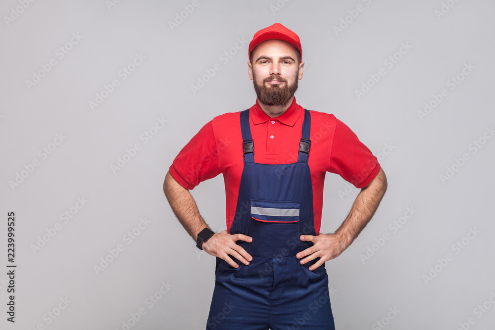Portrait of young confident repairman with beard in blue overall, red t-shirt and cap standing and holding hands on waist with smile, indoor, studio shot, isolated on gray background, copy space.