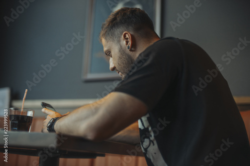 Young attractive pensive man looking at mobile phone screen and drinking soda.