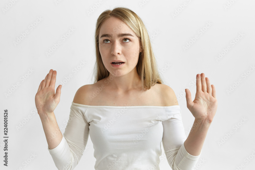 Horizontal studio shot of irritated annoyed young blonde female in stylish top rolling eyes and making stop gesture, being fed up with lies and excuses of her boyfriend who is late for dinner
