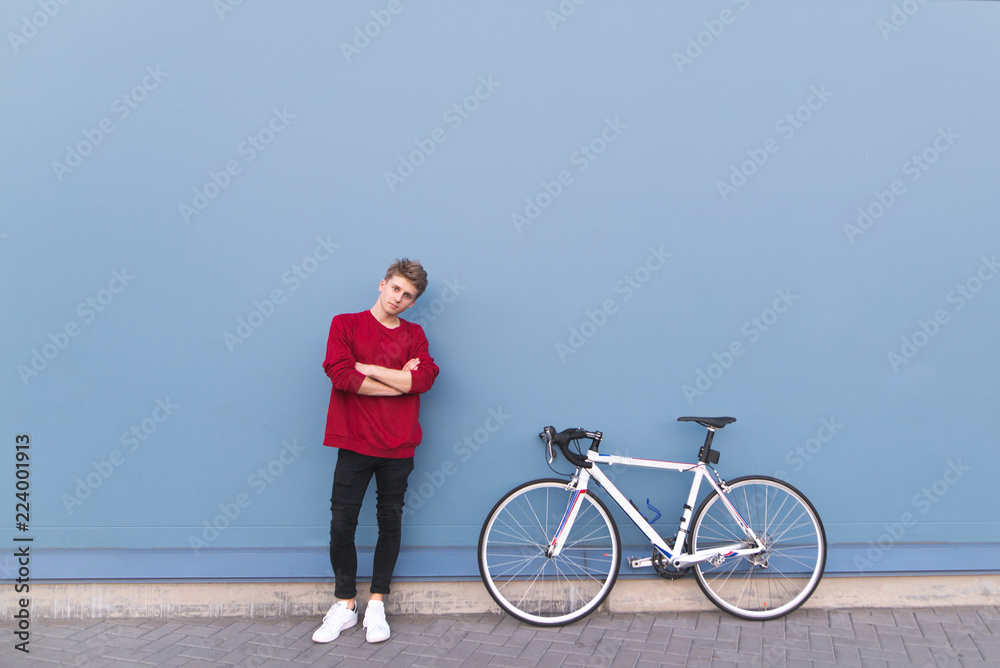 Fototapeta premium Attractive young man in a red sweatshirt standing by a bicycle on a blue background and looking at the camera. Portrait of a stylish student with a bike. Minimalism. Copyspace