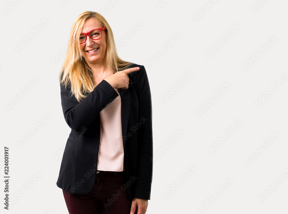 Middle-age blonde business woman with black jacket pointing to the side with a finger to present a product or an idea while looking forward smiling on isolated grey background Middle-age blonde business woman with black jacket pointing to the side with a finger to present a product or an idea while looking forward smiling on isolated grey background