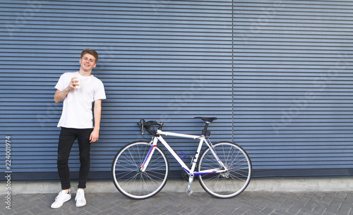 Portrait of a happy young man standing with a cup of coffee in his arms, near a bicycle on the background of the wall, looking in camera and smiling. Happy student with a bike looking into the camera.