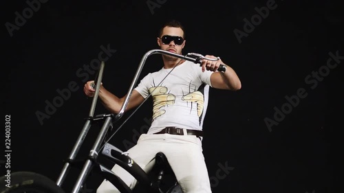 Brutal young bearded man with sunglasses dressed in white t-shirt and trousers with brown leather belt is sitting on black steel bicycle, gripping handles, moves backward and forward. photo