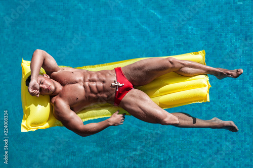 Muscular young sexy wet naked guy in a speedo lying on inflatable mattress in pool