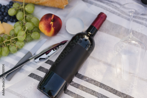 Mockup of a bottle of whine lying on a white cotton carpet during a picnic. Having lunch in a public park with red wine fruit, cheese and baguette. Copy space for you lwine logo, design or text