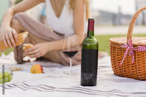 Close up of a blank bottle of red wine. Girl having picnic on the background. Space for you logo or design. Woman having lunch outdoors and enjoying holidays and weekends in the park with green grass